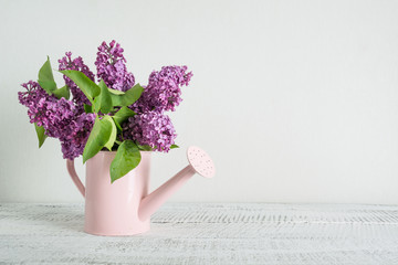 Decorative pink watering can with purple lilac flowers on white wooden table. Gardening concept. Spring and summer.