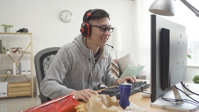 Excited male gamer playing winning online video game on personal computer in mess bedroom. Young asian man with handsome smile wearing earphones concentrated looking monitor. cozy afternoon home