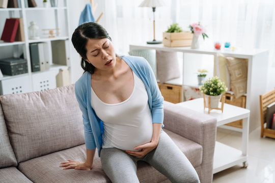beautiful asian pregnant woman with big belly baby and hands holding frowning face hard to stand up from couch sofa in living room. illness body ache lady try to sit down to relax release painful.