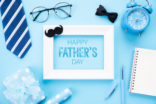 Happy fathers day concept. Top view of blue tie, beautiful gift box, alarm clock, white picture frame with Happy father's day text and retro film camera on bright blue pastel background. Flat lay.