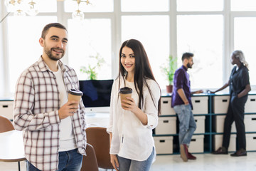Coffee break. Two young people holding coffee cups and discussing something with smile in front of collegues at office