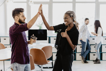 Celebrating success. Cheerful young indian man and african woman giving high-five while having coffee break in creative office