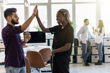 Celebrating success. Cheerful young indian man and african woman giving high-five while having coffee break in creative office