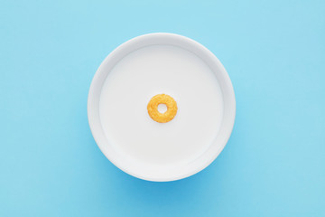 Single cereal ring floating on a white bowl full of milk on blue background. Close up top down view. - Powered by Adobe