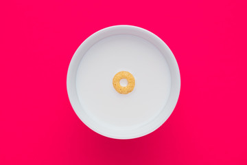 Single cereal ring floating on a white bowl full of milk on pink background. Close up top down view.