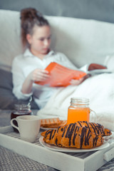 Young woman eating healthy breakfast in bed Soft cozy photo of a young teen girl on a bed with a cup of juice in her hands. A girl is sitting on a bedspread in a shirt and near a breakfast tray.