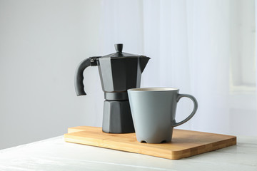 Cutting board with cup of fresh coffee and coffee maker on white table against light background, space for text