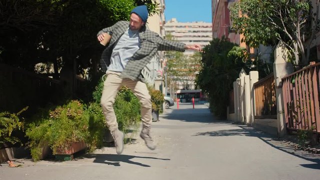 Cheerful trendy guy dances jumping around calm street enjoying happy life and new music, Hispanic dancer having funny leisure and walking at sunny green city landscape alone, dressed in blue hat