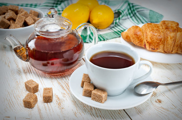 Still life white Cup with tea and transparent kettle and fresh croissant on a wooden light table.