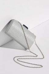 Front view shot of bright gray clutch, adorned with massive faceted golden clasp fastener. The accessory with snake-like placed chain strap is angled on the white platform. Trendy women's accessory.