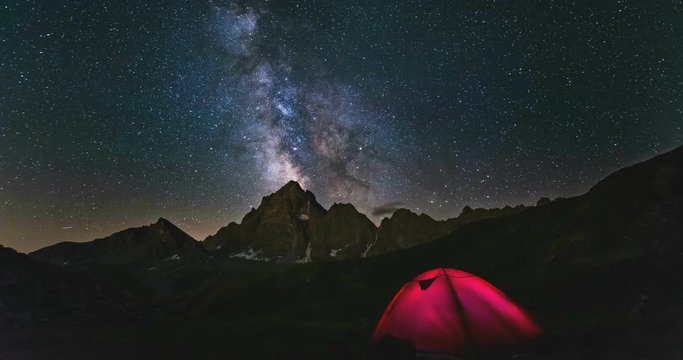 Time lapse Milky way galaxy stars rotating over the Alps, camping illuminated tent, snowcapped mountain peak, astro night sky stargazing