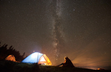 Night summer camping in the mountains. Couple travellers covered by blanket plaid sitting together beside campfire and tourist tent. On background forest and starry sky full of stars and Milky way.