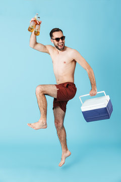 Full length photo of joyful shirtless man wearing sunglasses smiling while carrying cooler with cold beer