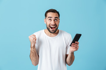 Photo of excited brunette man wearing basic t-shirt laughing and holding smartphone