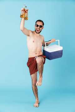 Full length photo of happy shirtless man wearing sunglasses smiling while carrying cooler with cold beer