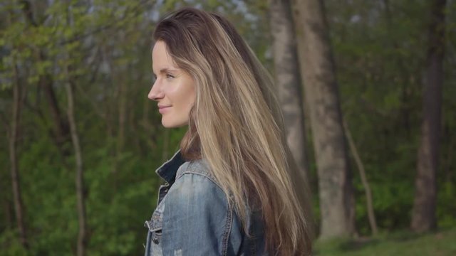 Portrait of a beautiful smiling young woman wearing a denim jacket walking on a beautiful sunny day outdoors. Charming girl with a beautiful smile, confident look and long beautiful hair. Slow motion.