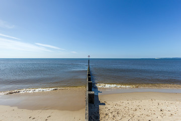 A view of a sandy beach with a groyne (breakwater) and calm sea under a majestic blue sky