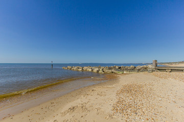 A view of a sandy beach with a stony groyne (breakwater) and calm crystal sea under a majestic blue sky