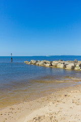 A view of a sandy beach with a stony groyne (breakwater), calm crystal sea and cormorant spreading his wings on the weathercock under a majestic blue sky