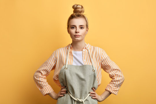 Young Serious Woman In Apron Posing With Arms On Hips And Looking At The Camera. Close Up Photo. Studio Shot. Housewife Is Ready To Work. People, Free Time, Hobby, Spare Time