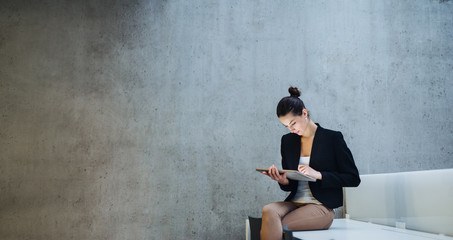 Young student or businesswoman sitting on desk in room in a library or office, using tablet.