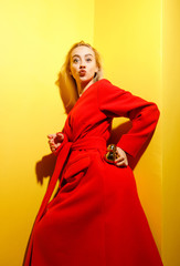 Fototapeta premium Fashion young girl blogger dressed in stylish red coat poses with the gold little duck figurine in her hands on the background of yellow walls in the show room