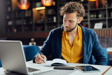 Young serious bearded Caucasian blogger dressed smart casual writing notes in agenda while sitting...