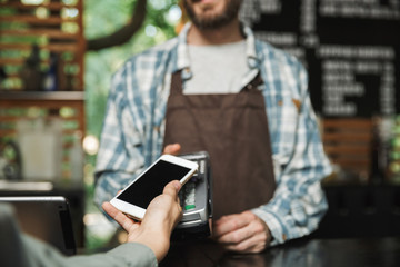 Portrait of caucasian barista man holding consumer terminal for paypass payment while working in street cafe or coffeehouse outdoor