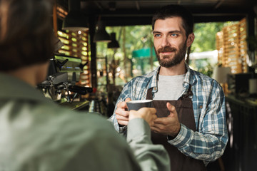 Portrait of unshaved barista man smiling while working in street cafe or coffeehouse outdoor