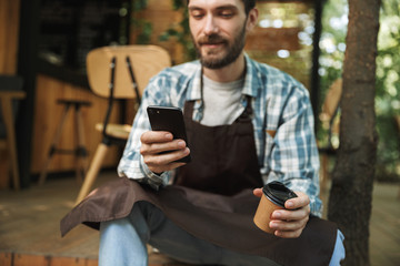 Photo of joyous waiter man using smartphone while working in cafe or coffeehouse outdoor