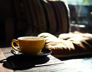 Hot coffee in a yellow ceramic mug placed on a wooden table along the glass wall with warm sunlight in the morning.