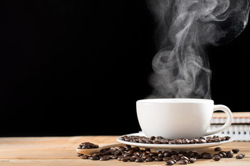 Hot coffee in a white coffee cup with hot steam Coffee break in the morning There are notebooks and roasted coffee beans placed on the plate and on the old wooden table. On a black background - images