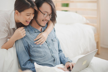 Young Asian man sit on the ground and working with laptop computer which woman lie on the bed behind man and they are smiling feeling happy in the bedroom at home. the life at home concept.