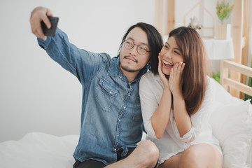 Young Asian man and woman are sitting on the bed and take a selfie photo which smiling feeling happy in the bedroom at home. the life at home, the couple love concept.