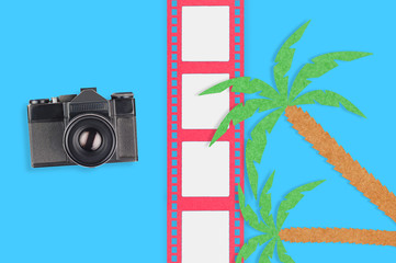 Old vintage camera for photo near palm trees and film strip cut out of colored paper on blue table. Top view. Travel concept
