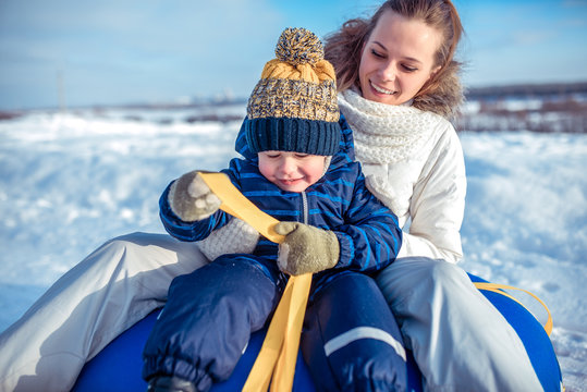 A young mother, woman with child, boy, son of 3 years old, in the winter outside in warm clothes, sitting on a tubing, rolling down a hill, playing, having fun, and relaxing on weekends in nature.