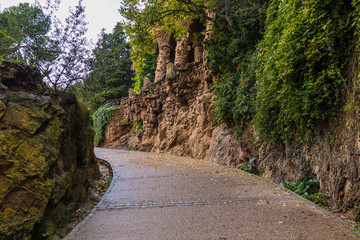 A footpath between two stone walls with lush vegetation in the Park Guell in overcast autumn day, Barcelona, Catalonia, Spain