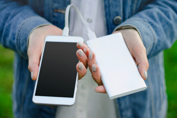 Girl holding power bank and a smart phone on green grass background. Girl charges her smartphone...