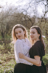 Fototapeta premium Portrait of two beautiful young sisters in the green spring garden, hugging in the field in the sunshine. Having fun together, positive emotions, bright colors. Copy space. Happy girlfriends at sunset