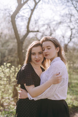 Fototapeta premium Portrait of two beautiful young sisters in the green spring garden, hugging in the field in the sunshine. Having fun together, positive emotions, bright colors. Copy space. Happy girlfriends at sunset