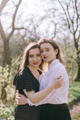 Fototapeta premium Portrait of two beautiful young sisters in the green spring garden, hugging in the field in the sunshine. Having fun together, positive emotions, bright colors. Copy space. Happy girlfriends at sunset
