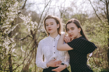 Fototapeta premium Portrait of two beautiful young sisters in the green spring garden, hugging in the field in the sunshine. Having fun together, positive emotions, bright colors. Copy space. Happy girlfriends at sunset