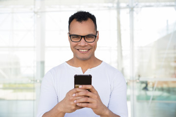 Medium shot of smiling young mixed-race man in spectacles and white T-shirt standing at office building, texting on phone. Front view. Lifestyle concept