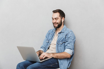 Photo of brunette laughing man having beard typing laptop and using earpod while sitting on chair