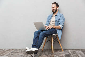 Photo of casual brunette man having beard typing laptop and using earpods while sitting on chair...