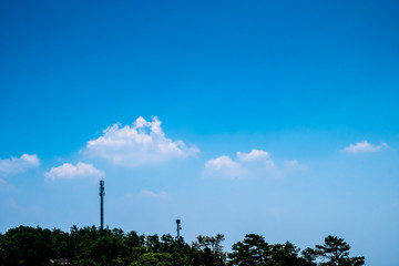 Green tree forest and telecommunication towers against clear blue sky with white fluffy clouds.