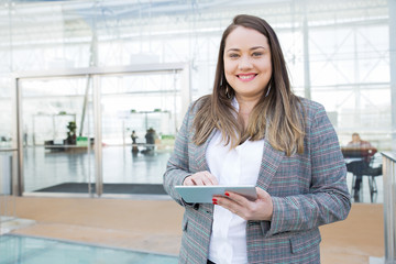 Positive lady with tablet posing in business center. Fat young woman in office jacket holding...
