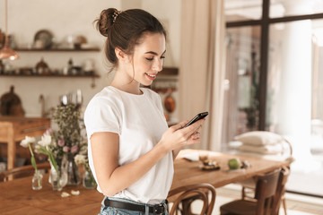 Image of optimistic nice woman typing on cellphone and smiling in cozy kitchen