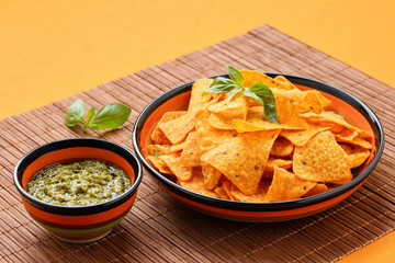 Nachos corn chips, basil pesto and leaves in colorful bowls on a bamboo mat and orange tablecloth. Copy space