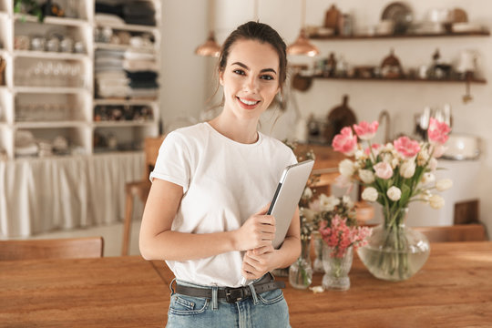 Image of nice european woman holding laptop computer and smiling at camera in cozy kitchen